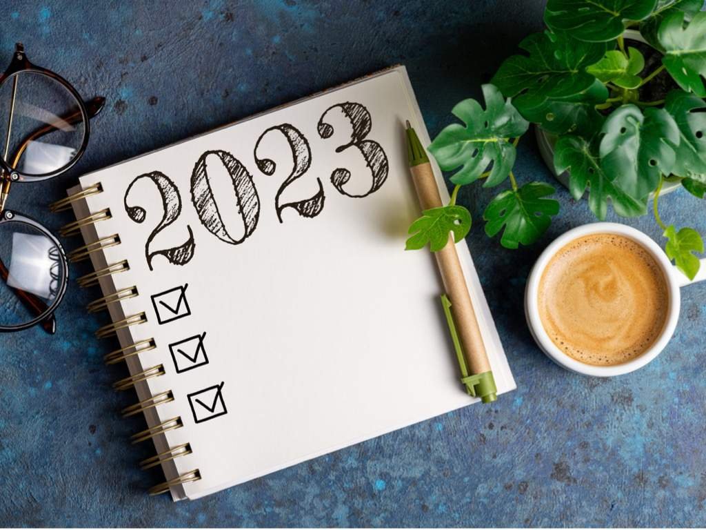 Notizbuch mit der handgeschriebenen Jahreszahl 2023 und drei abgehakten Kästchen, daneben Brille und Kaffee